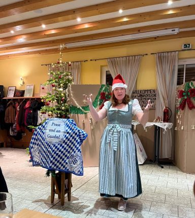 Frau in traditionellem Kleid mit Weihnachtsmütze vor einem geschmückten Baum.